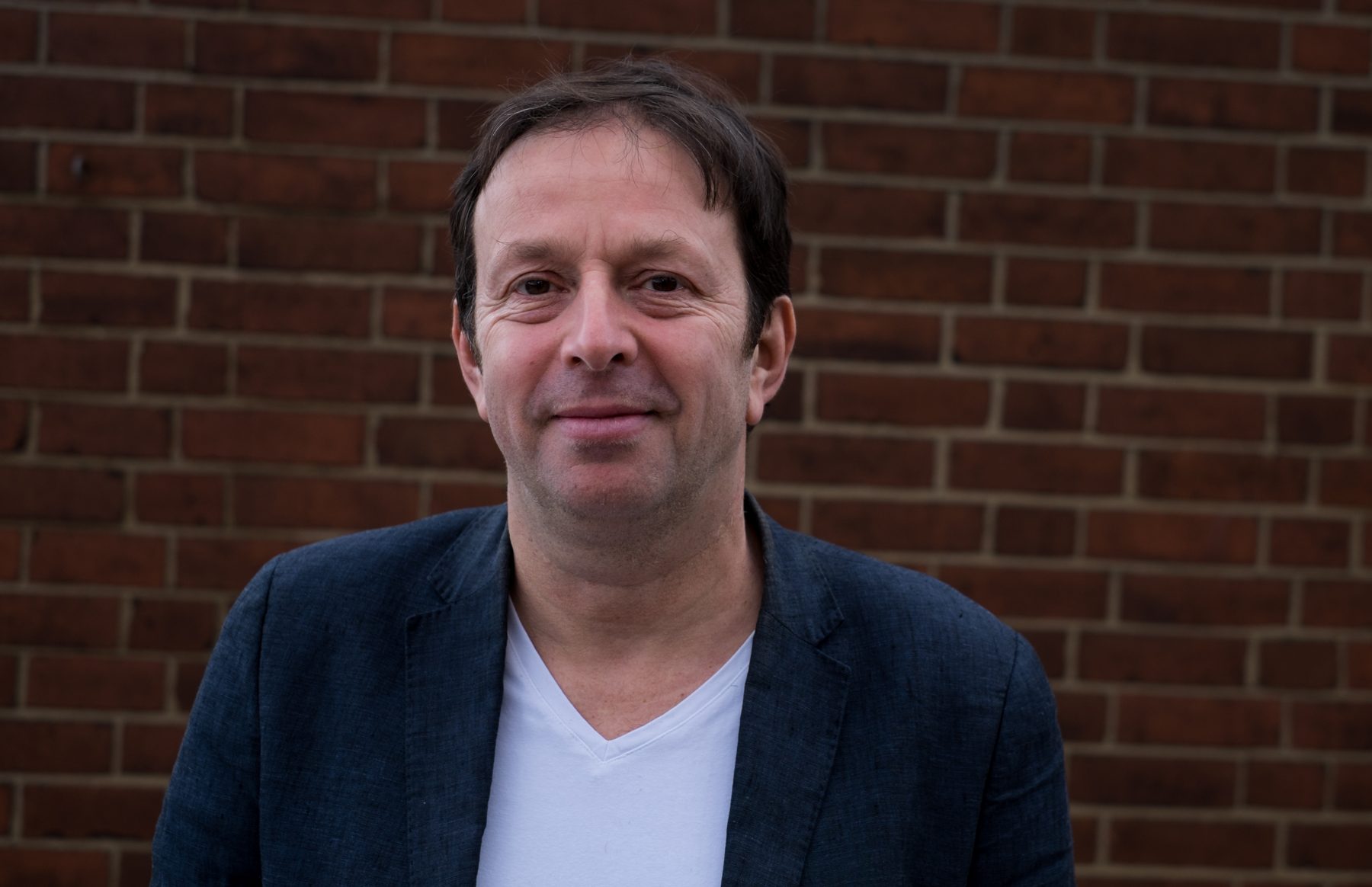 Mark Maier smiling in a navy blazer against a brick wall