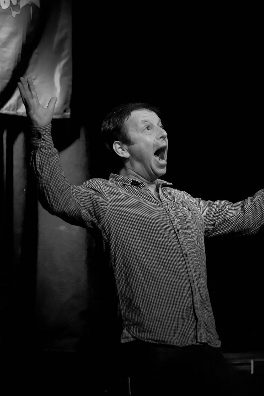 Black and white photo of Mark Maier on stage, arms flung wide, mid-punchline