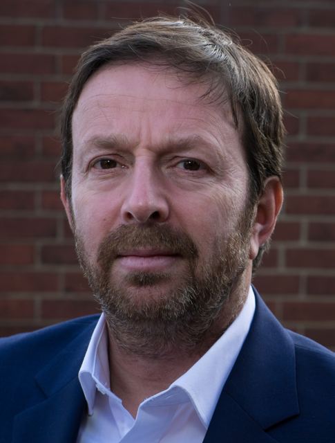 Head and shoulders portrait with beard, navy suit, brick wall